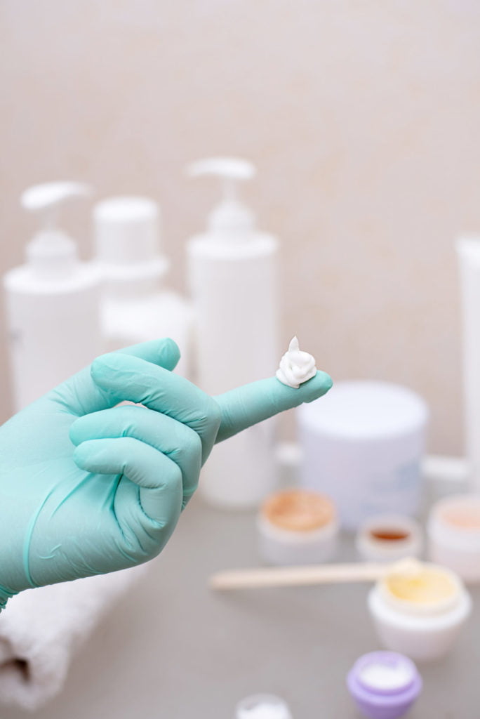 A drop of cream on a hand in a medical glove on the background of a table with jars of cream, skin care concept, close up.