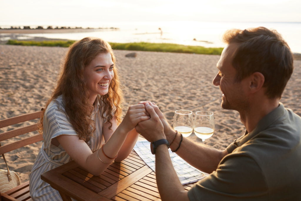 Portrait of happy young couple holding hands at picnic outdoors while enjoying romantic date on beach, copy space