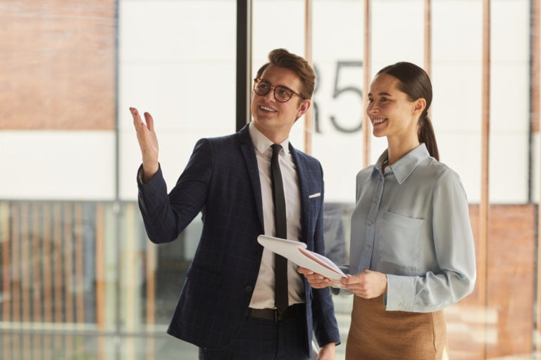 Waist up portrait of smiling real estate agent discussing property with female client and pointing up while standing in empty office building interior lit by sunlight, copy space