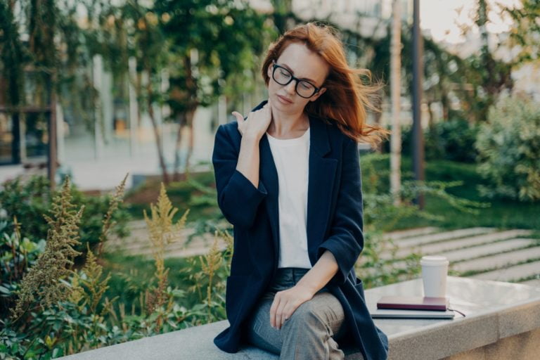 Young tired businesswoman massaging neck, feeling pain in body muscles because of bad posture at work, sitting in park after long working day in office, overworked female suffering from backache,