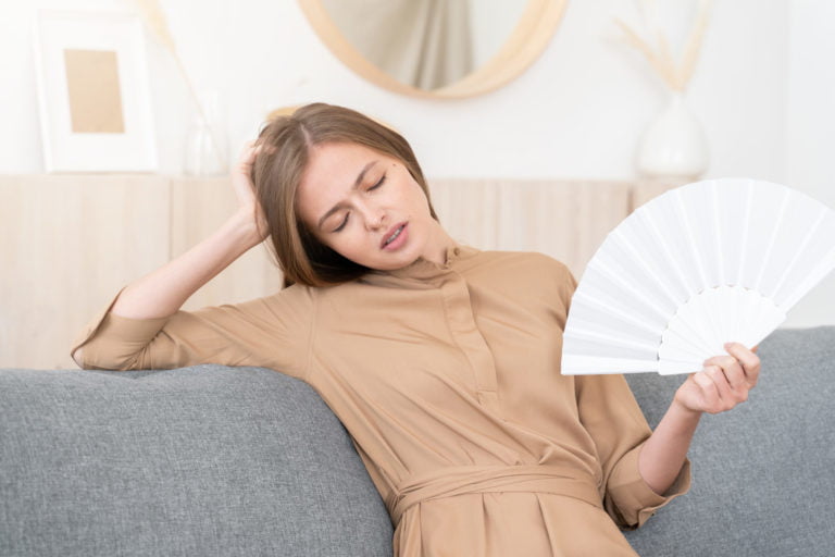 Young woman in beige dress suffering from heat at home with no air conditioner, using paper fan
